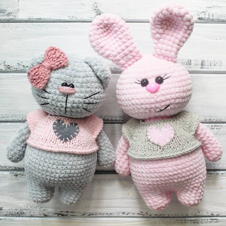 May include: Two crocheted stuffed animals, a gray cat with a pink bow and a pink bunny, both wearing pink and gray sweaters with heart-shaped patches.