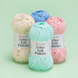 Puede incluir: Cinco ovillos de hilo YarnArt Cotton Gold Paillettes en crema, azul claro, verde menta y rosa. Cada ovillo tiene una etiqueta que dice "Cotton Gold Paillettes". El hilo está adornado con lentejuelas. Cada ovillo pesa 50g.