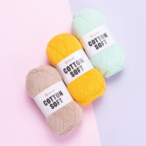 Pode incluir: Três novelos de fio YarnArt Cotton Soft em bege, amarelo e verde menta. Cada novelo tem uma etiqueta branca com o texto "COTTON SOFT". O fio está disposto sobre um fundo rosa e roxo.