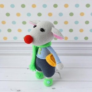 May include: A gray crocheted stuffed animal with a green scarf, blue shirt, and gray pants. The animal has a red nose, pink ears, and black eyes. It is holding a yellow crocheted object.