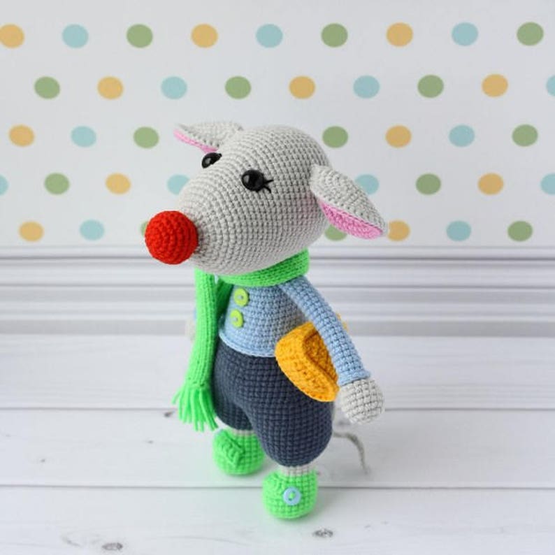 May include: A gray crocheted stuffed animal with a green scarf, blue shirt, and gray pants. The animal has a red nose, pink ears, and black eyes. It is holding a yellow crocheted object.