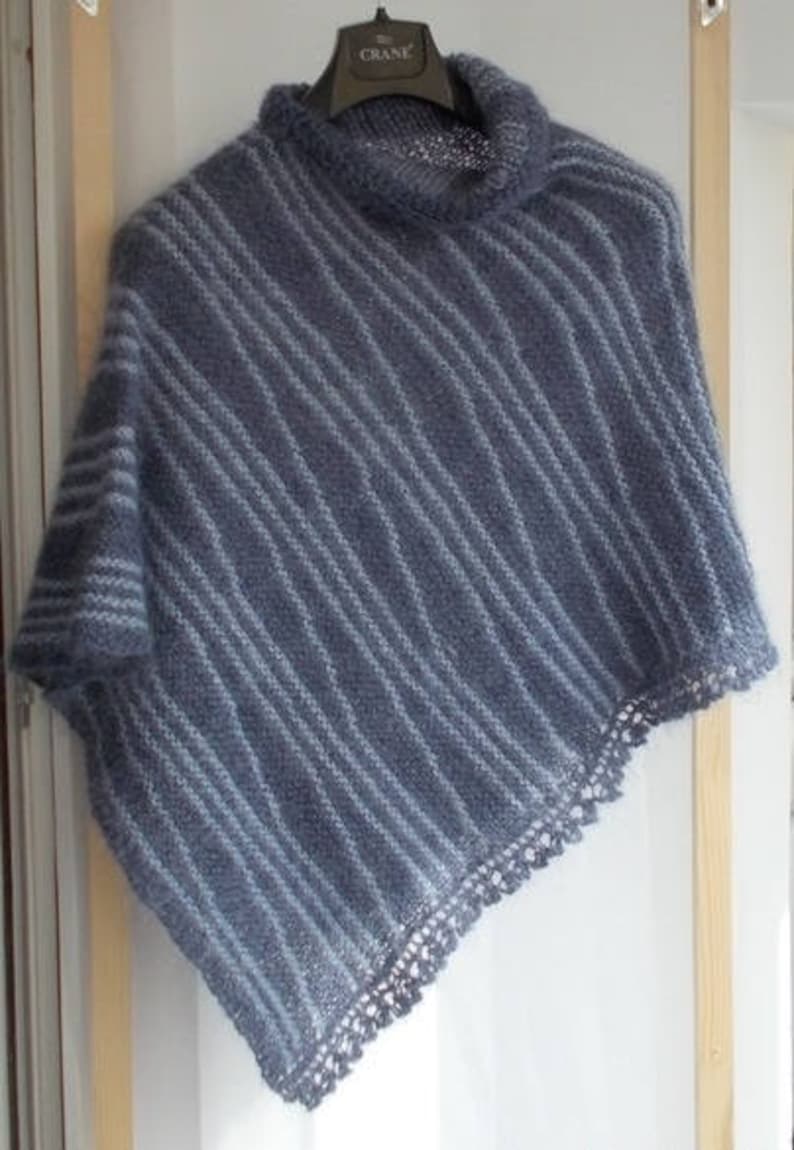 May include: A blue knitted poncho featuring a high cowl neckline and a diagonal striped pattern in lighter blue tones. The garment has a textured, fuzzy appearance and is finished with a delicate, openwork crocheted lace trim along the bottom edge.