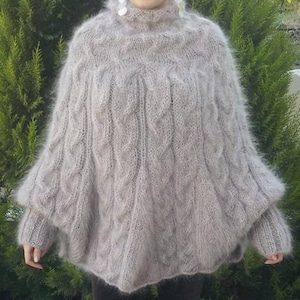 May include: A light beige, fuzzy, long-sleeved knit poncho featuring a high turtleneck collar and a prominent vertical cable knit pattern throughout the body. The garment has a loose, draped silhouette with ribbed cuffs at the ends of the sleeves.