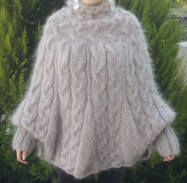 May include: A light beige, fuzzy, long-sleeved knit poncho featuring a high turtleneck collar and a prominent vertical cable knit pattern throughout the body. The garment has a loose, draped silhouette with ribbed cuffs at the ends of the sleeves.