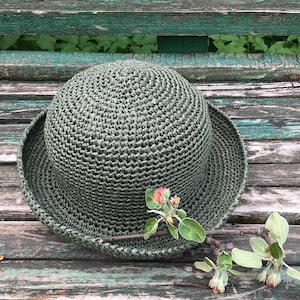 May include: A green crocheted hat with a wide brim, sitting on a weathered wooden surface with a small sprig of green leaves and pink blossoms.