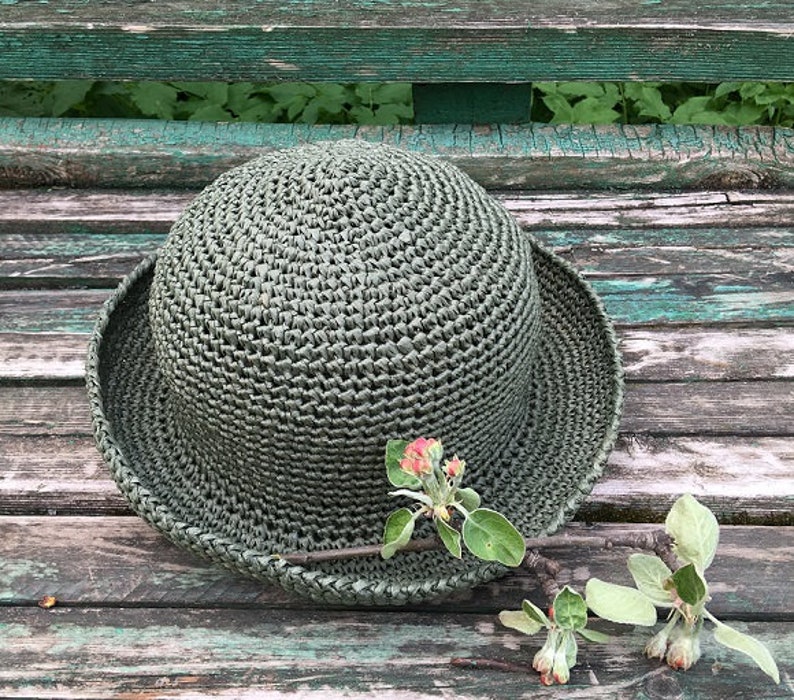 May include: A green crocheted hat with a wide brim, sitting on a weathered wooden surface with a small sprig of green leaves and pink blossoms.