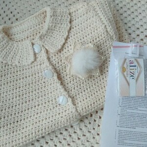 May include: A cream-colored crocheted baby cardigan with a white button closure and a fluffy white pom-pom detail. The cardigan is laid out on a cream-colored crocheted blanket. A small white card with the text "Alize Cotton Gold" is visible in the background.