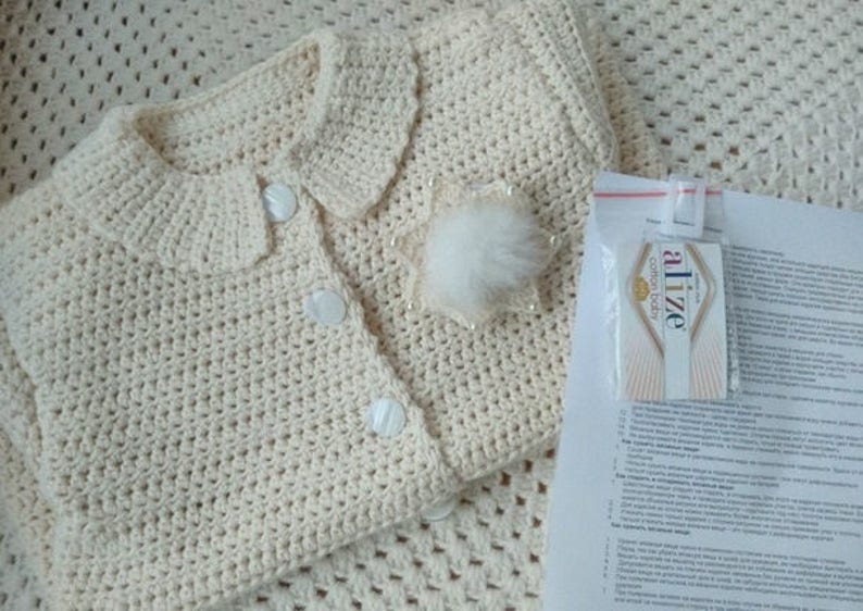 May include: A cream-colored crocheted baby cardigan with a white button closure and a fluffy white pom-pom detail. The cardigan is laid out on a cream-colored crocheted blanket. A small white card with the text "Alize Cotton Gold" is visible in the background.