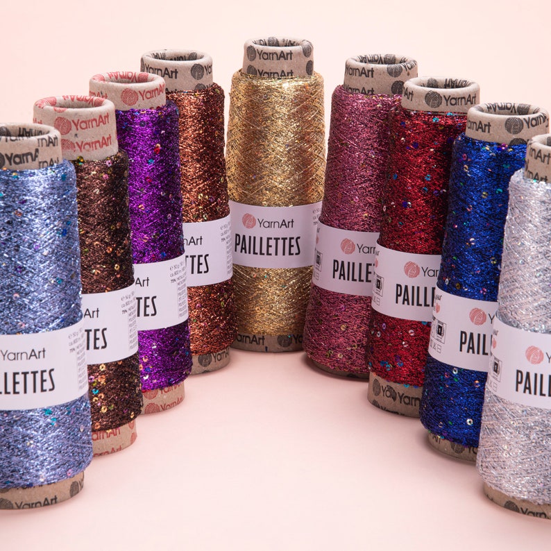 May include: A collection of spools of metallic yarn with sequins. The spools are arranged in a semi-circle on a pink background. The yarn is in various colors, including gold, silver, red, blue, purple, and brown. The spools are labeled with the words "YarnArt" and "Paillettes".