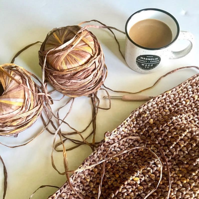 May include: Two balls of brown and yellow yarn with a crocheted piece of fabric and a mug with the text "Magnolia Market Waco TX" on a white surface.