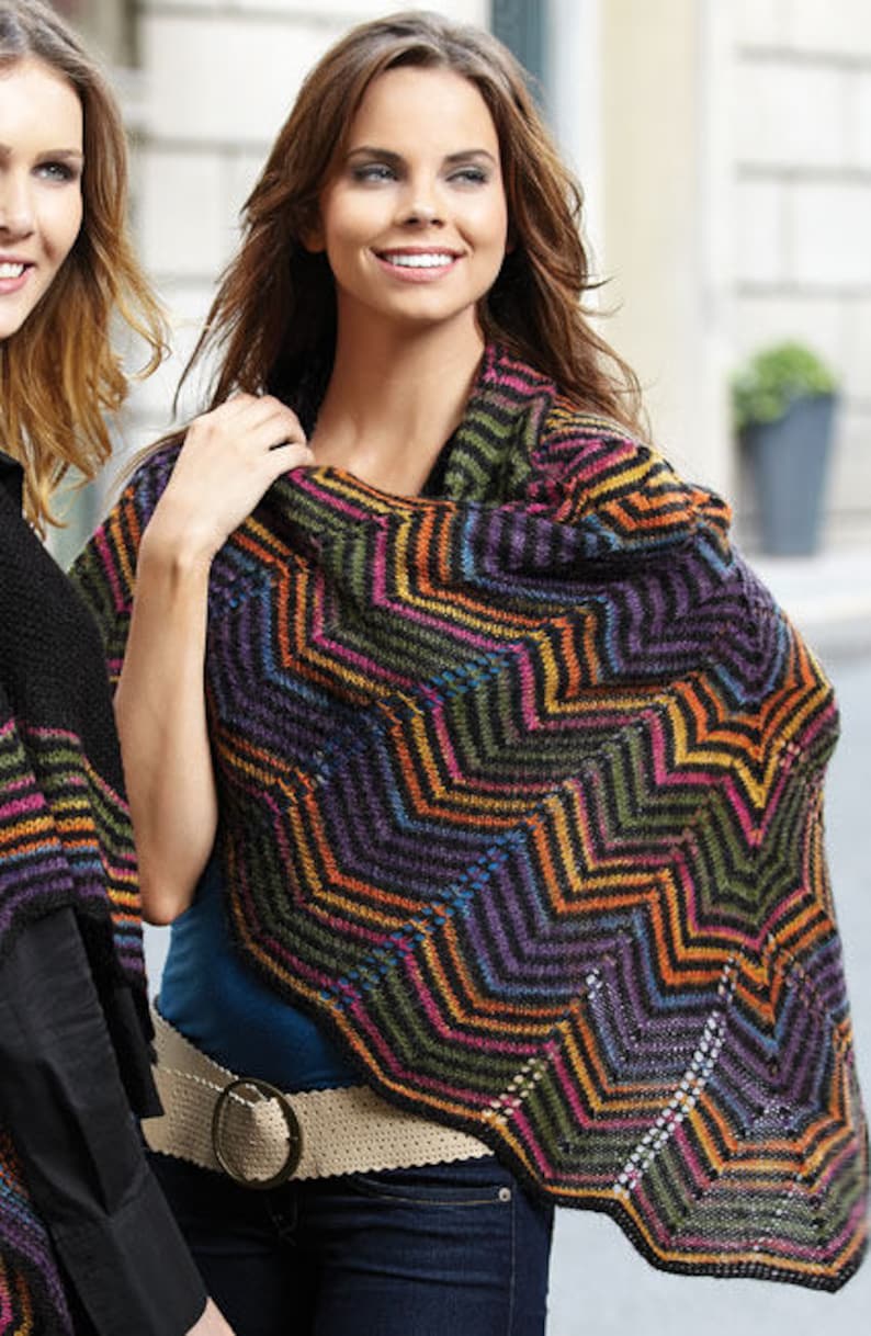Puede incluir: Un chal de punto colorido con un patr&oacute;n de chevron en tonos de negro, naranja, morado, verde y marr&oacute;n. El chal est&aacute; drapeado sobre los hombros de una persona que lleva pantalones vaqueros azules y un cintur&oacute;n marr&oacute;n.