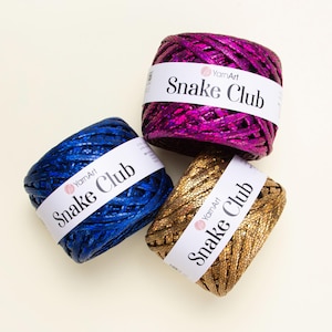 Op de afbeelding: Drie bollen YarnArt Snake Club garen in levendige kleuren: koningsblauw, fuchsia en goud. Elke bol heeft een textuur met een slangenpatroon en een wit etiket met de merk- en productnaam.