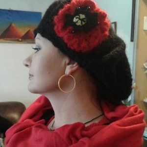 May include: A black beret with a red crocheted flower embellishment. The flower has a black center with a white design. The beret is worn by a person with a red scarf.