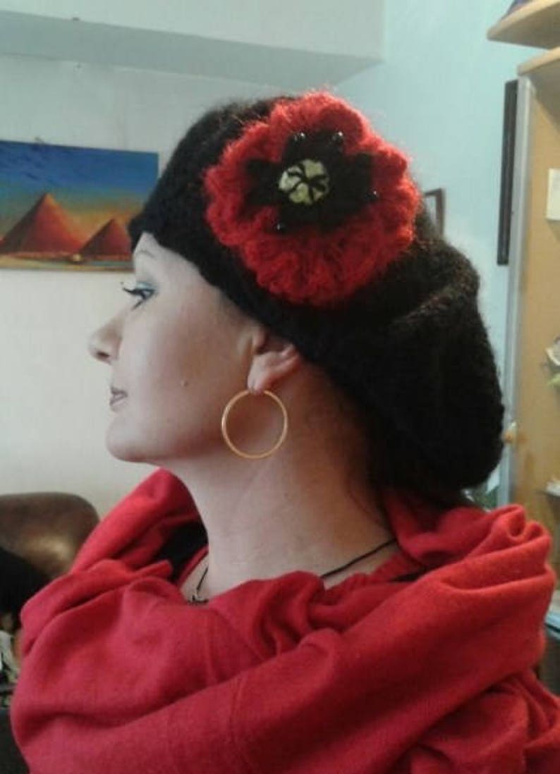 May include: A black beret with a red crocheted flower embellishment. The flower has a black center with a white design. The beret is worn by a person with a red scarf.