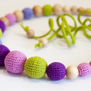Puede incluir: Un collar de cuentas de madera y crochet de colores con cuentas moradas, rosas, verdes y azul claro. El collar est&aacute; atado con un cord&oacute;n verde.