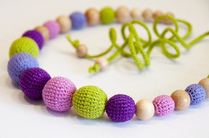 Puede incluir: Un collar de cuentas de madera y crochet de colores con cuentas moradas, rosas, verdes y azul claro. El collar est&aacute; atado con un cord&oacute;n verde.