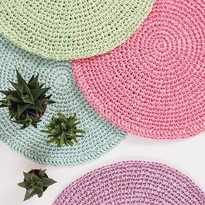 May include: Four crocheted coasters in different pastel colors: light green, pink, blue, and purple. The coasters are round and have a textured surface. Three small potted succulents are placed on the coasters.