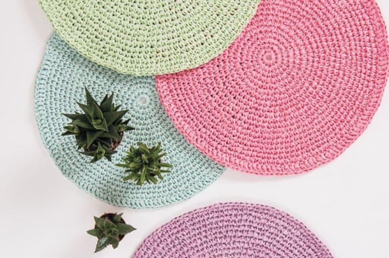 May include: Four crocheted coasters in different pastel colors: light green, pink, blue, and purple. The coasters are round and have a textured surface. Three small potted succulents are placed on the coasters.