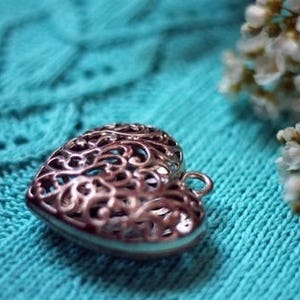May include: A silver heart-shaped pendant with a delicate filigree design, resting on a turquoise knitted fabric.