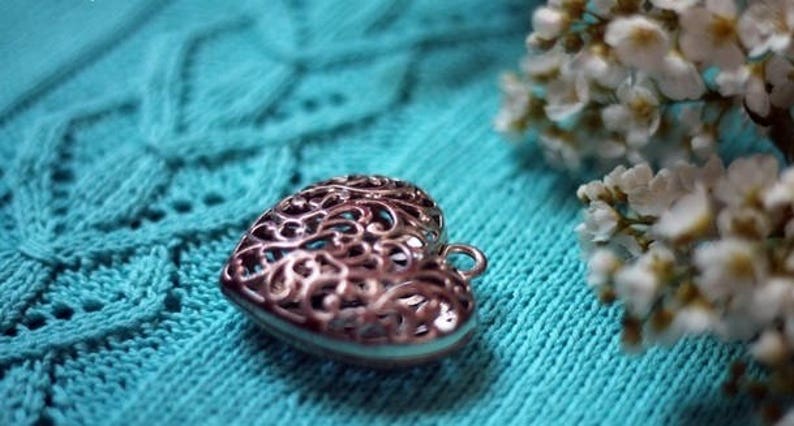 May include: A silver heart-shaped pendant with a delicate filigree design, resting on a turquoise knitted fabric.