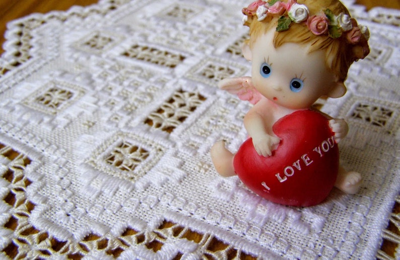 May include: A white porcelain cherub figurine holding a red heart with the words "I love you" on it. The cherub is wearing a flower crown and has wings. The figurine is sitting on a white crocheted doily.