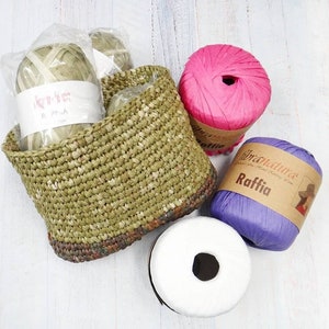 May include: A green crocheted basket with a brown trim, filled with four spools of raffia yarn. The yarn is in green, pink, purple, and white.