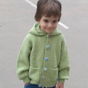 May include: A young child wearing a light green knitted cardigan with a hood and colorful buttons. The cardigan has two pockets on the front. The child is wearing blue jeans.