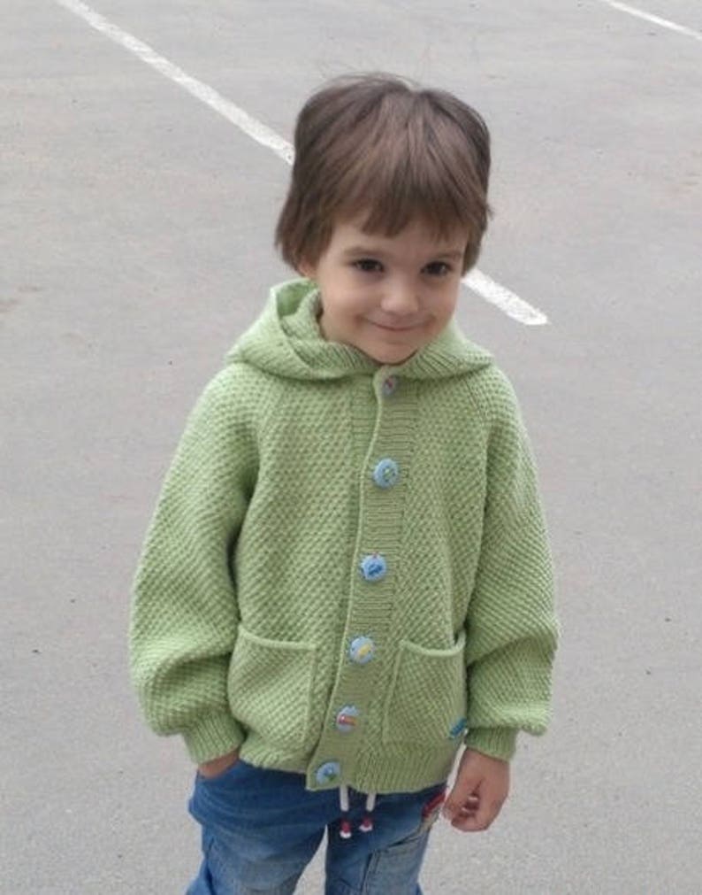 May include: A young child wearing a light green knitted cardigan with a hood and colorful buttons. The cardigan has two pockets on the front. The child is wearing blue jeans.