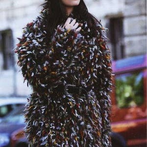 May include: A woman wearing a multi-colored fuzzy knit coat with a matching fuzzy knit hat. The coat has a black belt and the woman is wearing blue jeans.