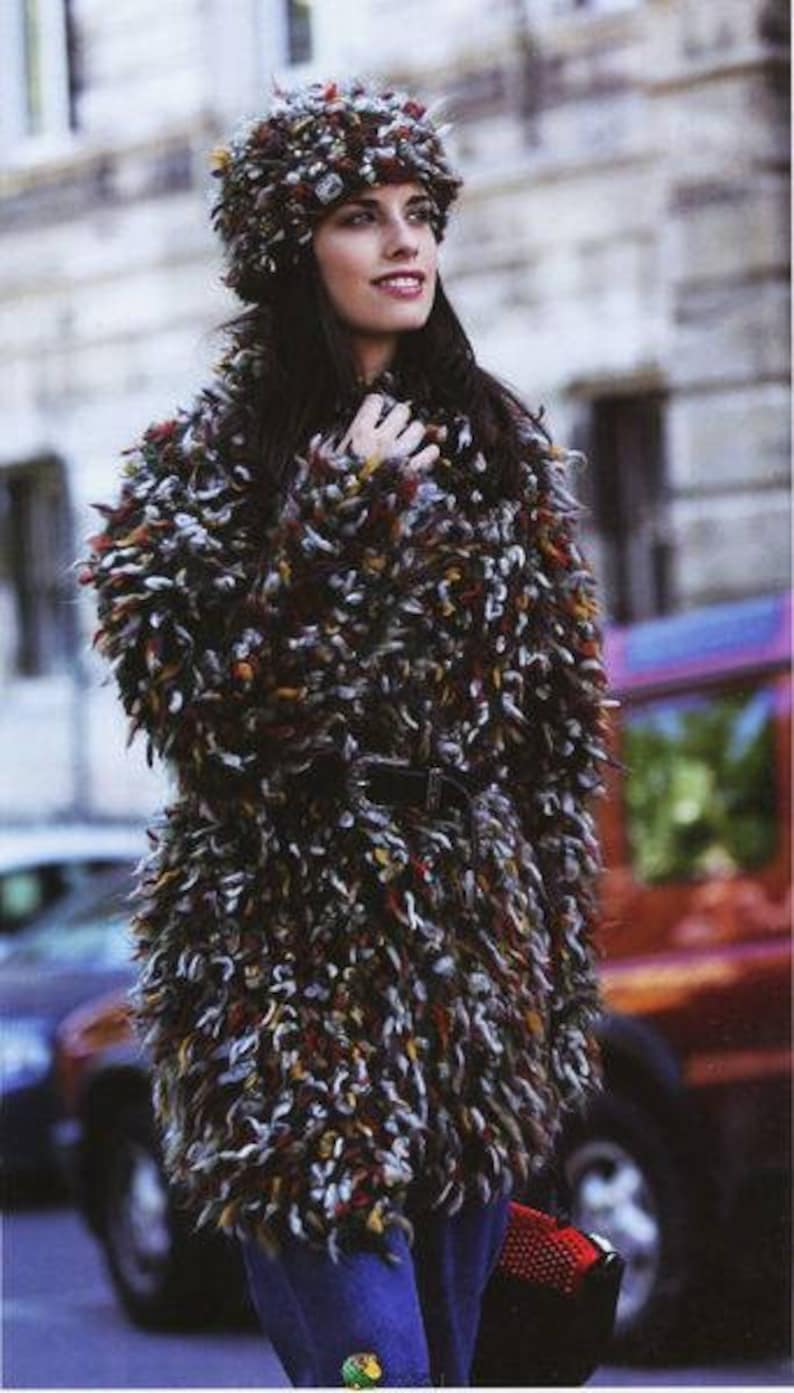 May include: A woman wearing a multi-colored fuzzy knit coat with a matching fuzzy knit hat. The coat has a black belt and the woman is wearing blue jeans.