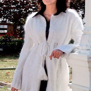 May include: A white, long-sleeved, fuzzy knit cardigan featuring a drawstring waist with tassel ends. The garment has an open front design, a round neckline, and a textured, soft appearance. It is worn over a solid black collared shirt.