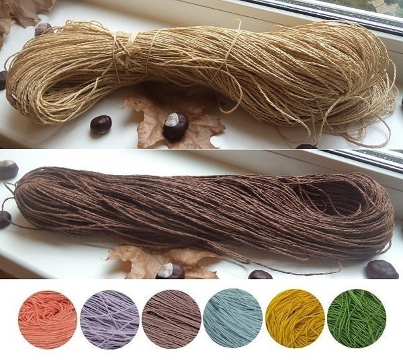Op de afbeelding: Twee spoelen natuurlijke vezel touw, een bruin en een beige. De spoelen zijn naast elkaar op een wit oppervlak geplaatst. Onder de spoelen bevinden zich negen kleine cirkels van gekleurd garen in tinten oranje, paars, bruin, blauw, geel en groen.