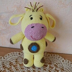 May include: A crocheted yellow giraffe toy with brown spots, pink nose, and blue eyes. The giraffe is standing on a crocheted doily.