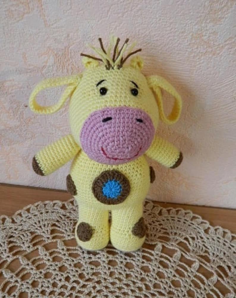 May include: A crocheted yellow giraffe toy with brown spots, pink nose, and blue eyes. The giraffe is standing on a crocheted doily.