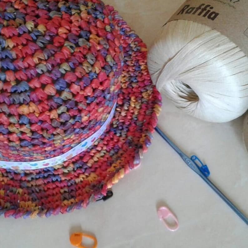 May include: A colorful crocheted hat with a white ribbon and a ball of raffia yarn. The yarn is labeled "Raffia".