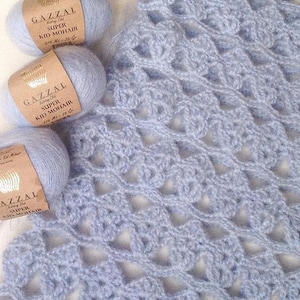 以下が含まれることがあります： 「GAZZAL Super Kid Mohair 375 ml - 25 g」とラベル付けされた薄い青色の糸の糸巻きが3つ、糸を使って作られたかぎ針編みの作品と一緒に写っています。かぎ針編みの作品は薄い青色で、オープンステッチのデザインです。