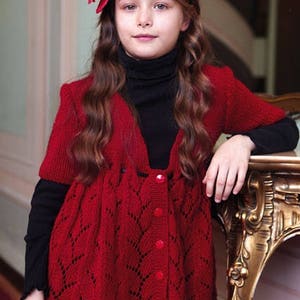 May include: A young girl wearing a red knitted cardigan with a button-up front and a delicate lace pattern. She is wearing a black turtleneck underneath and has a red flower headband in her hair.