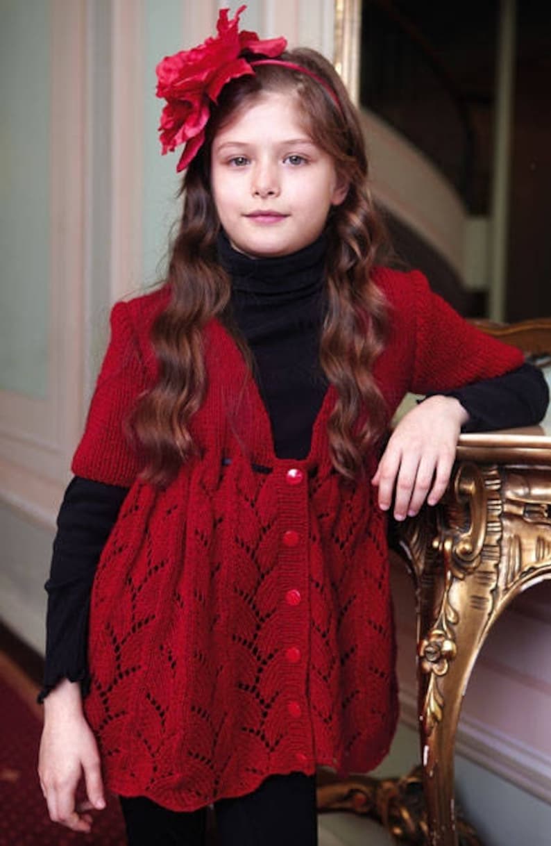 May include: A young girl wearing a red knitted cardigan with a button-up front and a delicate lace pattern. She is wearing a black turtleneck underneath and has a red flower headband in her hair.