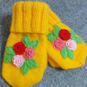 May include: A pair of yellow knitted mittens with crocheted flower embellishments. The flowers are in shades of pink, red, and green.