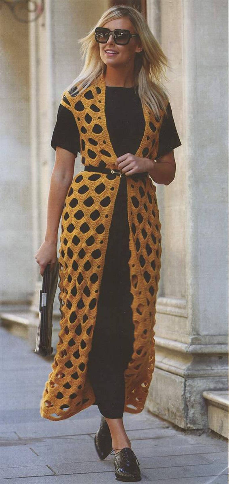 May include: A woman wearing a long, yellow crochet vest with a black top and black shoes. The vest has a geometric pattern and is open in the front. The woman is wearing sunglasses and a belt.
