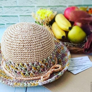 May include: A crocheted straw hat with a beige crown and a multi-colored brim. The brim is crocheted with a variety of colors, including pink, green, and yellow.