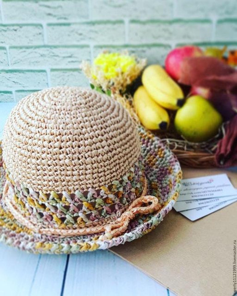 May include: A crocheted straw hat with a beige crown and a multi-colored brim. The brim is crocheted with a variety of colors, including pink, green, and yellow.