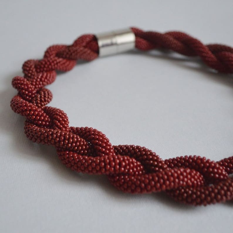 May include: A red beaded necklace with a silver clasp. The necklace is made of small, round beads that are woven together in a braided pattern.