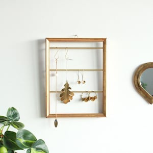 Pode incluir: Um organizador de joias dourado com uma moldura retangular e barras horizontais. Vários brincos dourados, um colar e uma pulseira são exibidos. Um espelho em forma de coração é visível à direita, e uma planta verde está no canto inferior esquerdo.