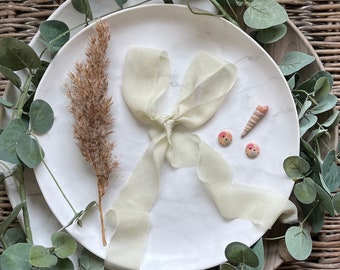 May include: A white marble plate with a light green silk ribbon tied in a bow. There are two small floral buttons and a seashell on the plate. A sprig of dried brown grass is on the left side of the plate.