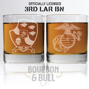 May include: Two clear glass whiskey tumblers with etched designs. The left tumbler features a skull and crossbones with the words "Wolf Pack". The right tumbler features the United States Marine Corps emblem. The text "Officially Licensed 3rd LAR BN" is above the tumblers. The text "Bourbon & Bull" is below the tumblers.