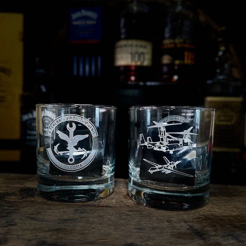 May include: Two clear glass tumblers with etched designs. The tumbler on the left has a circular design with a bird, anchor, wrench, and the text "A Tenebris Vindicta Venit 727 SOAMXS". The tumbler on the right has a design of a helicopter and a drone.