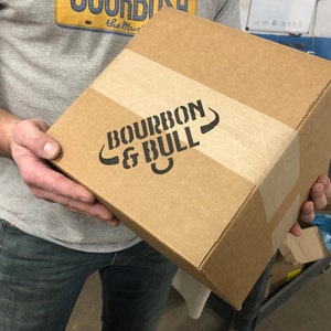 Puede incluir: Una caja de cart&oacute;n marr&oacute;n con las palabras "BOURBON & BULL" impresas en negro en el lateral. La caja est&aacute; cerrada con cinta adhesiva y tiene una etiqueta de papel marr&oacute;n.