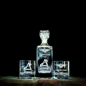 May include: A crystal decanter and two matching glasses with an engraved design. The decanter and glasses feature a winged emblem, the name "LT Thomas Logan 'TLO'", and the text "ARTEMIS OCT 01 JUNE 24".