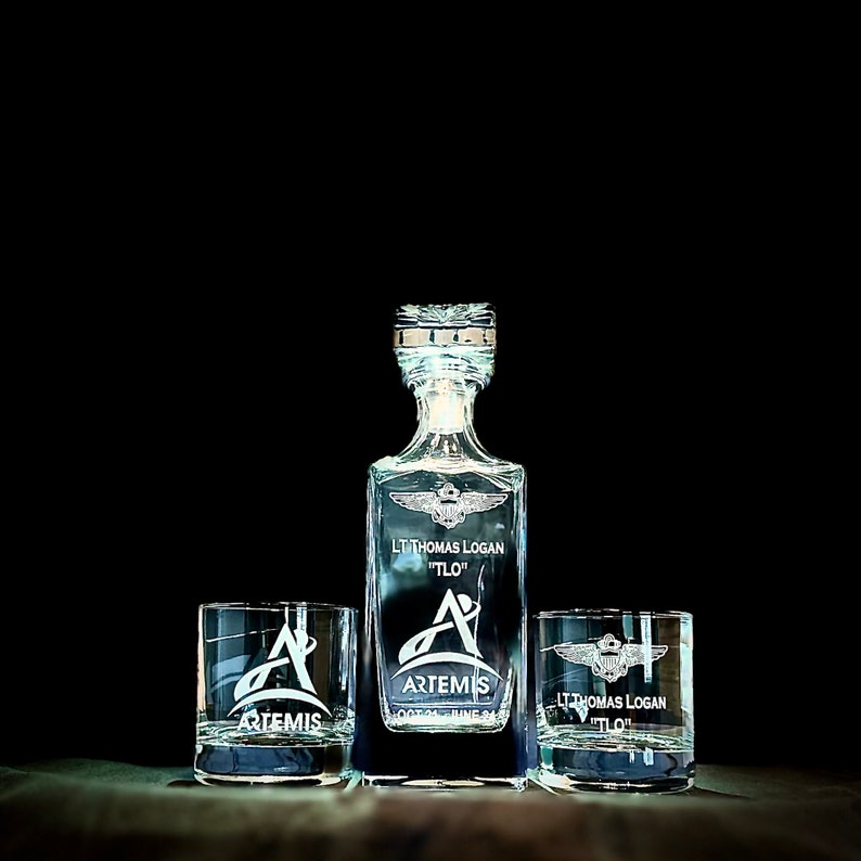 May include: A crystal decanter and two matching glasses with an engraved design. The decanter and glasses feature a winged emblem, the name "LT Thomas Logan 'TLO'", and the text "ARTEMIS OCT 01 JUNE 24".